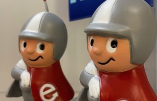 Zwei kleine Kunststofffiguren mit grauen Helmen und roten Mänteln mit weißen "e". Die Figuren sind Spardosen und stehen nebeneinander in einer Vitrine.