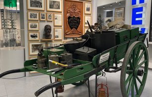 Eine einachsige grüne Schottsche Karre aus den 1930er bis 1960er Jahren steht vor einer Wand mit historischen Fotos des Stromversorgers. Auf der Karre befinden sich Ausrüstungsgegenstände der Monteure, darunter Zeltanlage, Hocker, Erste-Hilfe-Kasten, Kocher, Spitzhacken, Schaufeln, Spaten und Besen, die für Reparaturarbeiten an Erdkabeln und Freileitungen gebraucht wurden.