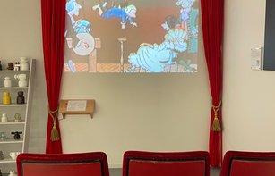 Unser kleines Museumskino mit sechs roten Sitzplätzen. Eine Filmprojektion läuft auf einer Wand, eingerahmt von roten Theatergardinen. Gezeigt werden kurze Filme elektrotechnischen Themen.